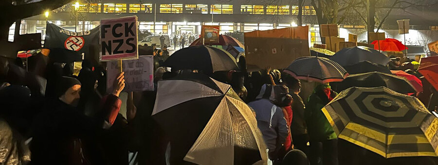 Eine große Demonstrantengruppe steht bei später Dämmerung vor der Oberpfalzhalle in Schwandorf mit Protestschildern und vielen Regenschirmen. Ein Schild hat die Aufschrift "FCK NZS". 