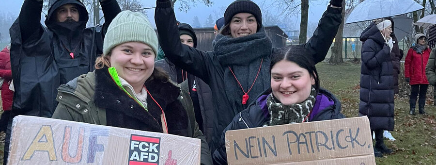 Eine Gruppe junger Menschen protestiert gegen die AfD und hält bunte, teils humorvolle Schilder hoch. Botschaften wie „Menschenrechte statt rechte Menschen“ und „Nein Patrick, die AfD ist keine Alternative“ sind zu lesen. Eine Frau mit grüner Mütze lächelt und hält ein regenbogenfarbenes Plakat mit der Aufschrift „Auf keinen Fall Digga“. Die Demonstrierenden tragen warme Kleidung und Pfeifen um den Hals. Im Hintergrund sind weitere Protestierende mit Schildern sowie Regenschirme zu sehen. Die Atmosphäre wirkt entschlossen und kreativ.