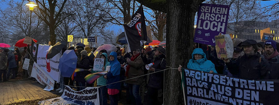 Eine große Gruppe Demonstrierender protestiert am Abend gegen die AfD und Rechtsextremismus. Sie halten Schilder, Banner und Fahnen mit Aufschriften wie „Rechtsextreme raus aus den Parlamenten!“ und „Sei kein Arschloch“. Viele tragen Regenkleidung und Kapuzen, da es nass ist. Mehrere Menschen haben Megafone, andere halten Regenschirme oder bunte Flaggen. Im Hintergrund sind Bäume, Straßenlaternen und das Schwandorfer Landratsamt mit erleuchteten Fenstern zu sehen. Die Stimmung ist entschlossen und kämpferisch.