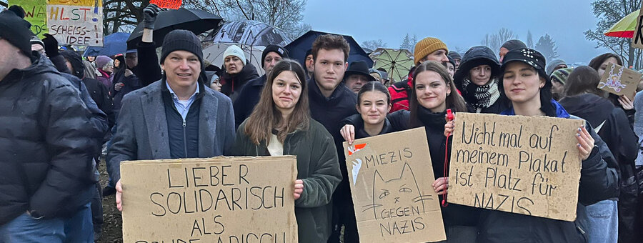 Eine Gruppe von Demonstrierenden steht eng beieinander und hält Protestschilder gegen Rechtsextremismus. Die Schilder tragen Botschaften wie „Lieber solidarisch als solide arisch“ und „Nicht mal auf meinem Plakat ist Platz für Nazis“. Viele Teilnehmende tragen Winterkleidung, einige halten Regenschirme. Im Hintergrund sind weitere Demonstrierende mit Plakaten zu sehen. Die Stimmung wirkt entschlossen und solidarisch.