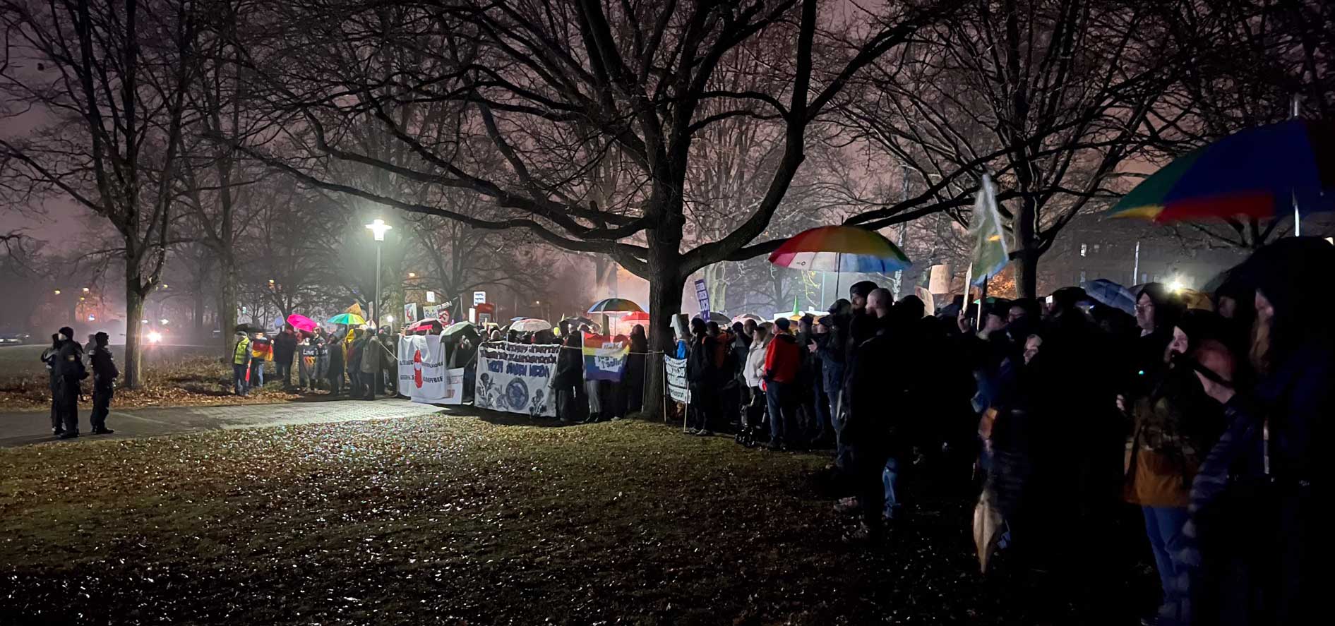 Eine große breit aufgestellte Menscheansammlung steht demonstrierend bei später Dämmerung im Park vor der Oberpfalzhalle in Schwandorf. Es werden Transparente und Schilder gegen Rechtsextremismus und gegen AfD gezeigt. Es regnet und viele halten Regenschirme. Mit etwas Abstand steht eine kleine Gruppe der Einsatzkräfte der Polizei. 